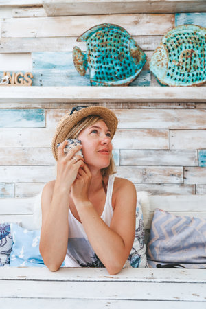 Portrait of a woman in a white tank top, straw hat, and sunglasses, holding a seashell to her ear as though she is listening to the ocean. She is sitting in the cafe with a sea vibeの写真素材