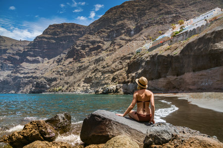 Enjoying the scenic view at Los Gigantes Beach, Tenerife, a woman in swimwear and a straw hat sits on a rock by the water, capturing the essence of a tropical vacationの写真素材