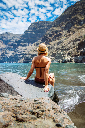 A woman in a swimsuit and straw hat sitting on a rock at the water's edge of Los Gigantes Beach, Tenerife, a scenic travel destinationの写真素材