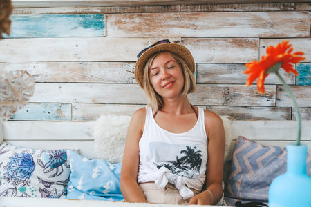 A happy woman with closed eyes relaxing in a cafe with a sea vibe, rustic wooden wall behind her, capturing a sense of coziness and casual comfortの写真素材