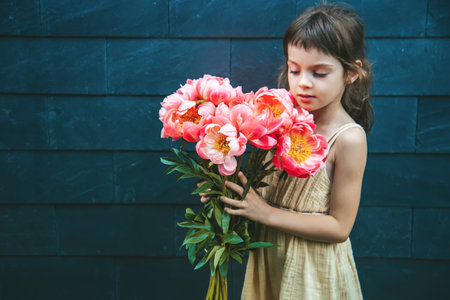 A beautiful little girl holding a large bouquet of pink peonies in front of a black background, highlighting the vibrant flowersの写真素材