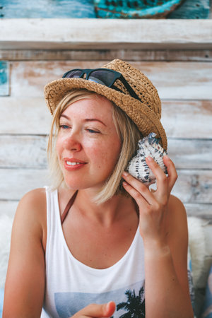 A portrait of a woman wearing a white tank top and a straw hat adorned with sunglasses, holding a seashell close to her ear, as if listening to its soundの写真素材