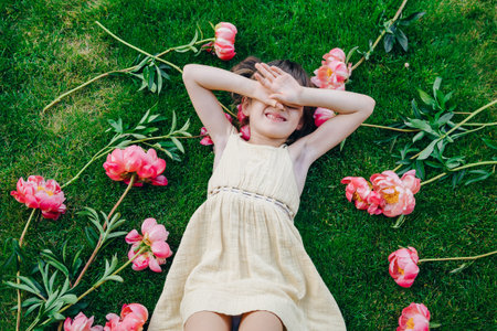 Little girl wearing a yellow dress, lying on the grass with pink peonies around her, capturing the striking contrast between the green grass and the bright pink flowers in a tranquil settingの写真素材