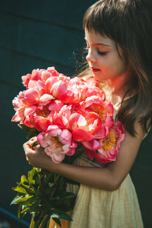 Beautiful little girl holding a large bouquet of pink peonies, illuminated by the sun, showcasing the striking flowersの写真素材