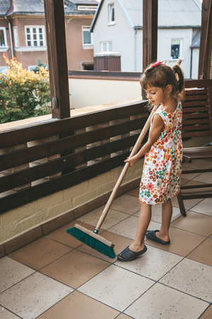 A little girl 5-6 years old is sweeping an outdoor balcony with a large broom, with wooden railings and residential buildings in the backgroundの写真素材