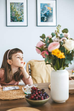 A little girl 5-6 years old dreaming or thinking about something, with flowers in a white vase on a foreground, and a bowl of grapes and cherriesの写真素材