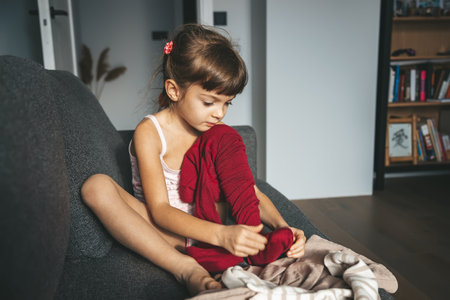A cute little girl sitting on a sofa in a room with a bookshelf, pulling on burgundy tights, getting dressed in the morning. Home life and morning routine.の写真素材