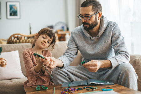 In a well-lit living room, a father and his 5-6 year old daughter sit on a couch, crafting with small construction pieces at a wooden tableの写真素材
