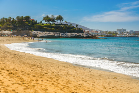 The serene shores of Playa Del Duque in Tenerife reveal calm, crystal-clear waves washing up against the soft sand. With vibrant greenery in the background, this idyllic beach is perfect for a peaceful holiday or relaxation contentの写真素材