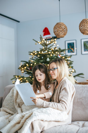 A mother and her little daughter nestled on the couch under a soft blanket, reading together, with decorated Christmas tree on the background. An inviting setting perfect for seasonal contentの写真素材