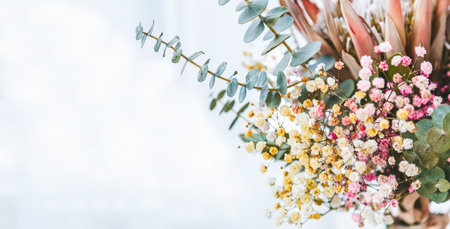 A close-up view of a floral arrangement against a blurred white background. The arrangement consists of small pink and yellow flowers with prominent green leaves and eucalyptus branches.の写真素材