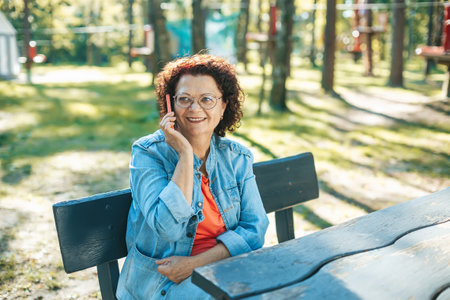 A smiling woman in her 60s sitting comfortably on a park bench, talking on her phone. She's wearing a denim jacket, with green trees and a calm park environment framing the scene.の写真素材