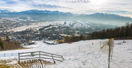 Snowy slopes in Zakopane, Poland, teem with skiers and snowboarders, descending towards a valley lined with scattered buildings. The snow-capped mountain range in the background under a cloudy sky creates a perfect winter setting for winter sports and alpine explorationの写真素材
