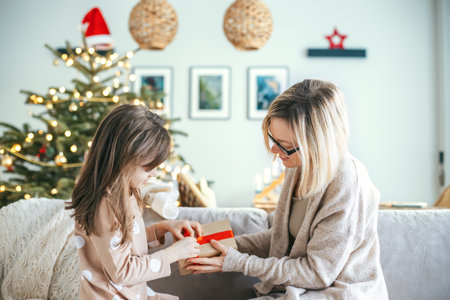 A woman and her daughter are seated on a couch, a girl is opening a holiday gift, as a Christmas tree adorned with ornaments and lights glows warmly in the backgroundの写真素材