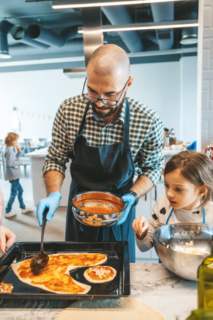 Father and daughter pouring sauce onto pizza dough, while making a pizza, sharing a fun and...