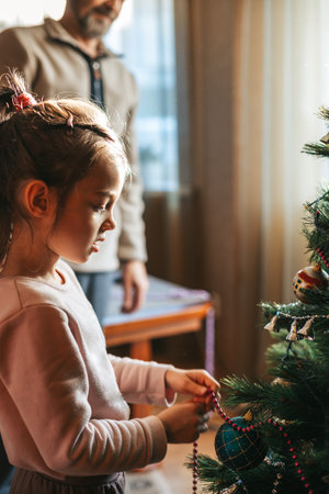A little girl decorating a Christmas tree with a string of beads. Her grandfather standing in the background observing the child.の写真素材