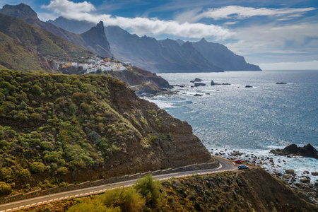 Scenic coastal landscape in Tenerife with winding road along rugged coastline, lush green hills, and dramatic mountain peaks partially covered by cloudsの写真素材