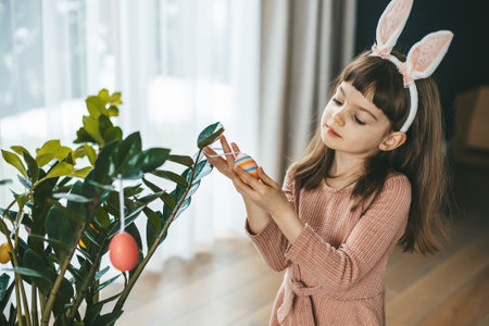 A sweet child in a pastel pink dress and bunny ears standing in a cozy indoor setting, adding decorative Easter eggs to a plantの写真素材