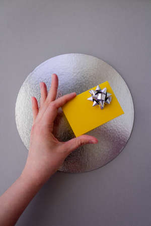 Woman hand holding yellow gift box with silver bow on grey background with a silver circle compositional center. Flat lay style. Festive greeting concept. Vertical viewの写真素材
