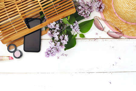 Set of summer women's accessories: bamboo bag, pink sunglasses, straw hat, cellphone, cosmetics and bouquet of lilac on white wood background. Space for textの写真素材