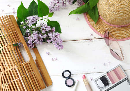 Set of summer women's accessories: bamboo bag, pink sunglasses, straw hat, cosmetics and bouquet of lilac on white wood background. Space for textの写真素材