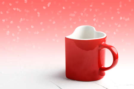 Red mug in shape of heart on white table at light red background with bokeh, front view. Valentine day concept background, copy space.の写真素材