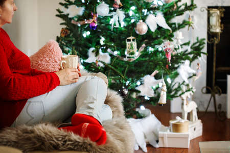 Woman sitting on a sofa with cup of hot beverage and looking on Christmas tree.の写真素材