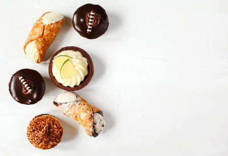Different pastries on white background: cannoli with ricotta, tartlets filled with citrus cream, fudge cake, tiramisu cake. Top view, copy spaceの写真素材