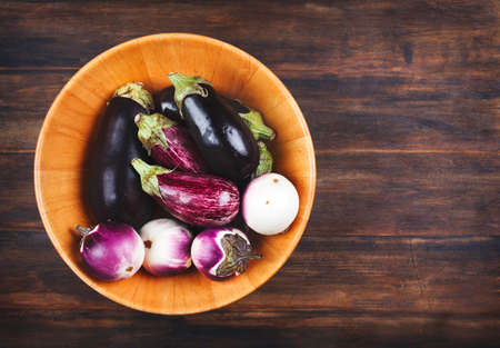 Fresh eggplants of different color and variety in wooden bowl on a wooden background, copy spaceの写真素材