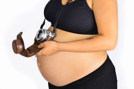 An analog camera with a brown leather casing hanging from a woman's neck by a strap and resting on her pregnancy bump.の写真素材