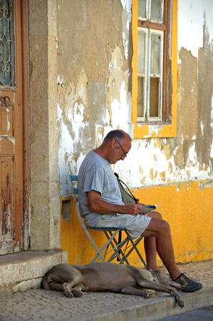 Algarve, Portugal, 24-September-2007: A man sits in a chair reading a newspaper with his dog beside him on a street in Algarve.のeditorial素材