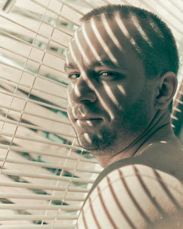 Handsom man standing at a window blinds and looking over his shoulder backの写真素材