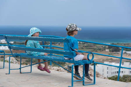 Small boy and girl sitting on a blue bench and looking down the hill on the seaの写真素材