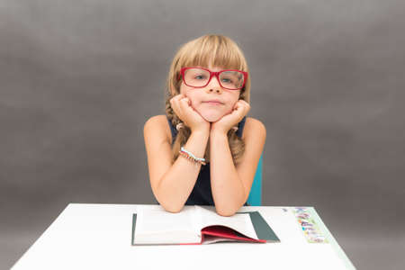 Young schoolgirl wearing big red glasses dreaming at a book with head leaning on his handsの写真素材