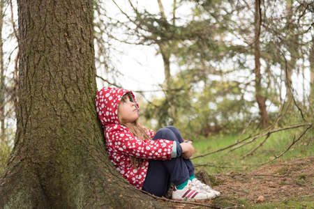 Little girl wearing red white jacket with a hood and sitting on a ground and leaning against a treeの写真素材