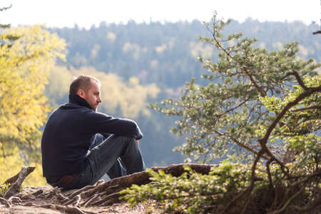 Handsome young man with beard sitting and contemplating on a rock in mountains. View  from his back with distant view blurred.の写真素材