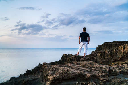 Young man standing on a sandstone rocks at the sea with hands in his shorts pockets and looking at horizonの写真素材