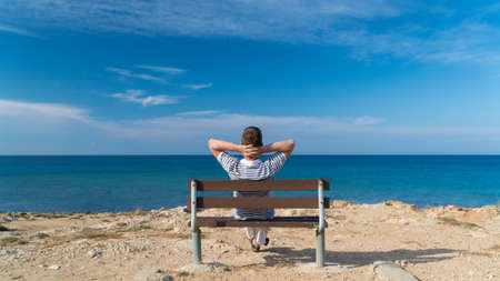 Young man sitting relaxed on a bench at a sea with his hands behind head and enjoying the viewの写真素材