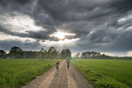 Dramatic cloudy sky with rays of sun light over mother with little girl cycling on an unmade road...