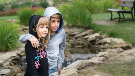 Young boy hugging his sister shoulder in a park. Both children wearing hoods and smiling.の写真素材