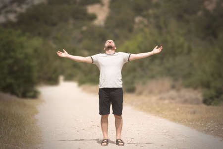 Young handsom man with beard standing on a path with his arms spread, head up and eyes closedの写真素材