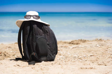 Sun glasses on white summer hat lying on top of a black backpack standing on a sandy beach at the seaの写真素材
