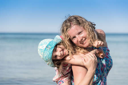 Young mother holding her little doughter on her back with sea in backgroundの写真素材