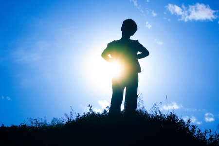Silhouette of a boy standing on a small hill with sun shining behind himの写真素材