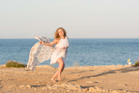 Young beautiful blond woman wearing white short dress dancing on a rocky sea shore with long scarfの写真素材