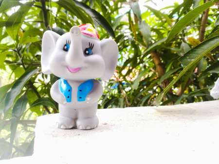 A Toy Elephant wearing a blue coat and a hat with long trunk and a smiling face.It is placed in a green background and a white floor.The two eyes are open and standing in both legs and other to body.の写真素材