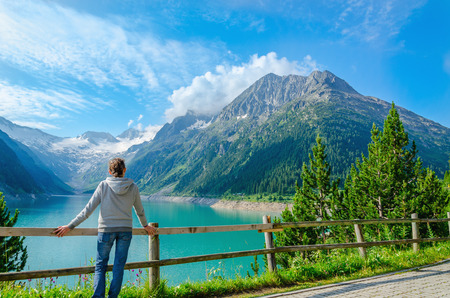 A young tourist stands beside an azure mountain lake on the background of the high peaks of the Alps, Zillertal, Austriaの写真素材