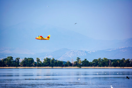 A yellow firefighting plane takes off from the surface of Lake Kerkini in Greece, with mountains and...