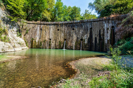 A scenic stone dam with small waterfalls flowing into a calm pond in Spilaio Kataraktes near Sidirokastro, Greece, surrounded by natural greenery.の写真素材