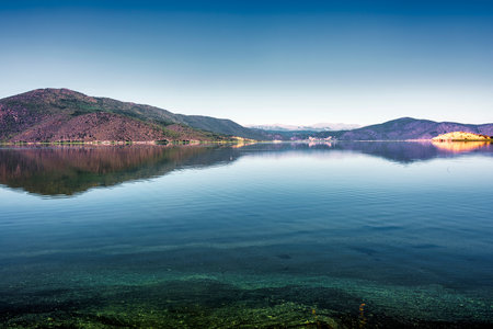 Clear reflections of lush green mountains on the tranquil waters of Lake Prespa under a bright blue sky in Mikrolimni.の写真素材